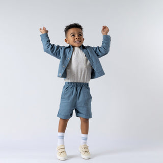 Boy smiling with arms in the air, wearing slate blue elasticated chino shorts, an ecru t-shirt tucked in, and a matching blue shacket, neutral background.