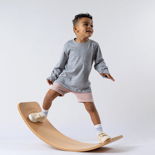 Young boy wearing a kids long sleeve t-shirt with stars, standing on a wooden wobble board and focusing on balancing. The star-print top in grey contrasts with dusty pink Aneby shorts, set against a neutral background.