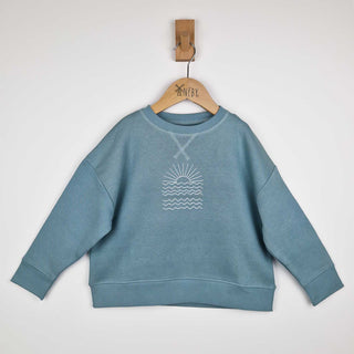 Flat lay of a steel blue kids sunrise sweatshirt of a white line drawn graphic sunrise above several rows of waves, displayed against a neutral background.