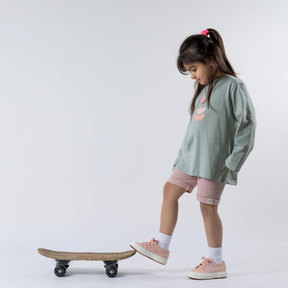 Girl wearing a children's sage green long sleeve t-shirt and dusty pink shorts, standing with one foot on a skateboard. She looks down slightly, set against a neutral background that highlights the relaxed fit and soft green tone.