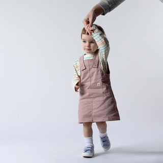 A young girl wearing a dusty pink organic cotton dungaree dress, standing against a neutral background. The dress features adjustable straps and a front pocket, paired with a soft long-sleeve top.