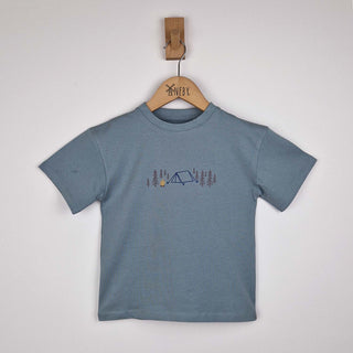 A kids' camping t-shirt laid flat, showcasing the front design. The print features a pitched tent, campfire, and fir trees on soft organic cotton fabric.
