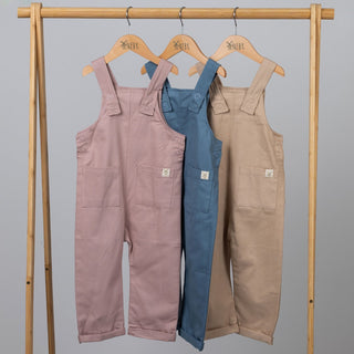 Product image displaying a set of three kids dungarees with patch pockets in blue, beige, and pink. The garments are neatly arranged on a wooden clothes rail on wooden hangers, highlighting their patch pocket details and fabric texture.