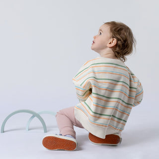 Back view of a small girl wearing a green striped sweatshirt, looking into the distance. Two wooden arch toys are visible against a neutral background.
