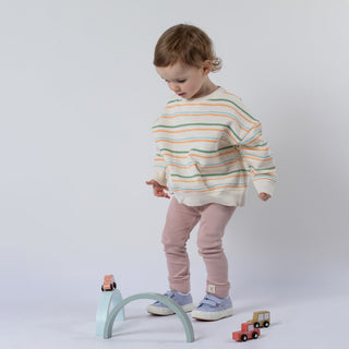 Small girl wearing a green striped sweatshirt on an ecru base with dusty pink leggings against a neutral background. She is looking down at small toy cars and pastel coloured wooden shapes.