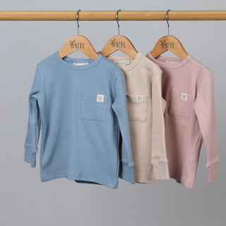 Three kids' long sleeve ribbed t-shirts in dusty pink, oat, and slate blue, hanging on wooden hangers from a wooden rail.