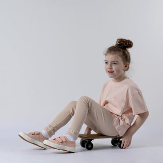 An older child wearing oat-coloured leggings sits on a small wooden skateboard. They are dressed in a blush pink t-shirt and are smiling while looking off into the distance. Their hands rest on the skateboard for balance.