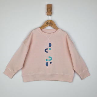 Flat lay of a kids pink sweatshirt featuring a unique design of vertical moons, horseshoes, and small suns in blue and green tones.