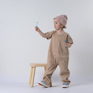 Small girl holding a bubble wand, wearing an oat short-sleeve overall with a pink beanie, standing in front of a wooden stool and neutral background.