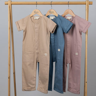 Three short-sleeve overalls in pink, blue, and oat, hanging on a wooden frame against a neutral background.