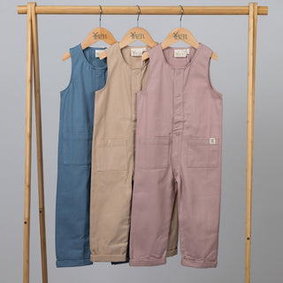 Three kids sleeveless overalls in dusty pink, slate blue, and oat, displayed on a wooden clothes frame against a neutral background.