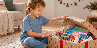 A boy in a blue camping-print T-shirt opens a Christmas present filled with colourful art supplies while sitting on a living room rug.