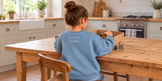A young child in a jumper sits at a wooden kitchen table, arranging toy animals in a cardboard box to make a little forest.