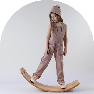 Girl standing on wobble board looking down, wearing pink sleeveless jumpsuit and matching pink beanie hat