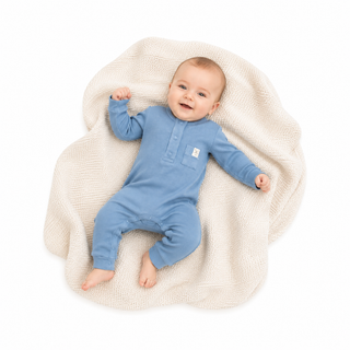 A baby lying on a textured cream blanket, wearing a light blue long-sleeve footless romper with a small white brand patch on the chest.