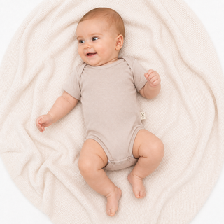 A baby lying on a cream-colored textured blanket, wearing a beige short-sleeve bodysuit. The baby is photographed from a top-down angle against a clean white background.