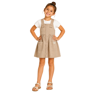 A young girl with her hair in a bun stands with hands on hips wearing a beige linen-look pinafore dress over a white t-shirt and tan sandals, photographed against a solid white background.