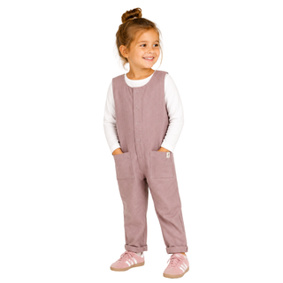 A young girl with her hair in a bun stands against a white studio background wearing a dusty rose corduroy overall jumpsuit over a white long-sleeve shirt, paired with white socks and matching pink trainers.