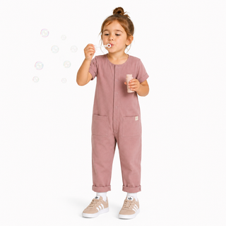 A young child wearing a pink short-sleeve jumpsuit with front pockets, standing against a white background and blowing bubbles while holding a bubble bottle.