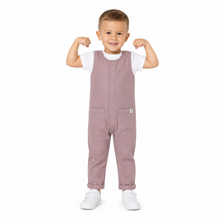 A young boy modelling a pink overall jumpsuit over a white t-shirt and white trainers, posing with his arms flexed against a solid white background.