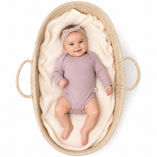 A baby wearing a pink long-sleeve bodysuit and a matching bow headband, lying on a cream knit blanket inside a woven basket with rope handles, photographed from above on a white background.