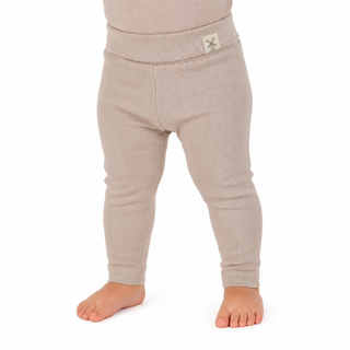 A close-up of a child's lower body wearing ribbed knit leggings in a light oatmeal beige color, standing barefoot against a white background.