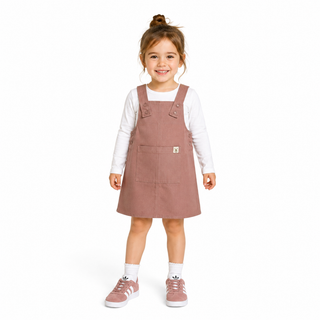 A young girl with her hair in a bun stands against a white background wearing a dusty rose corduroy pinafore dress over a white long-sleeve shirt, white socks, and pink trainers.