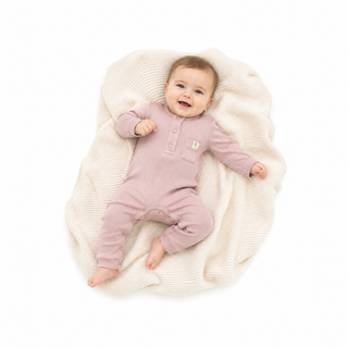 A baby lying on a cream ribbed blanket, wearing a dusty pink long-sleeve footless romper with a small embroidered logo on the chest.