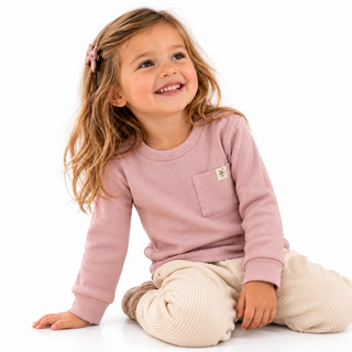 A young girl with light brown hair sits smiling on a white background, wearing a dusty pink long-sleeve crewneck sweatshirt with a chest pocket and cream-colored ribbed pants.