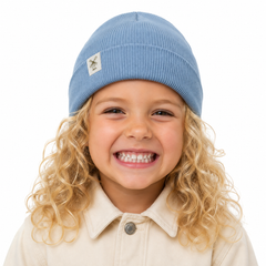 A smiling young girl with curly blonde hair wearing a light blue ribbed knit beanie and a cream button-up shirt, photographed against a solid white background.