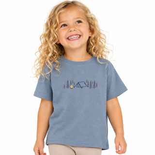 A young girl with curly blonde hair smiling while wearing a gre/blue short-sleeve t-shirt with a camping graphic on the chest, set against a plain white background.