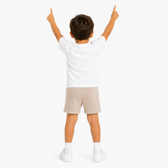 A child viewed from behind wearing a white short-sleeve t-shirt, beige shorts, white socks, and white trainers, standing against a plain white background with both hands raised.