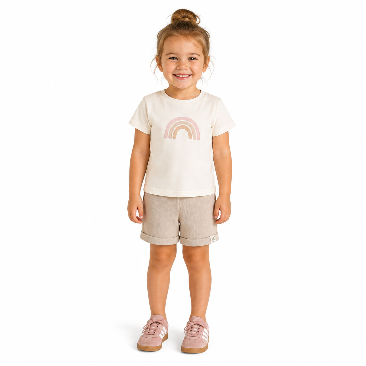 A young girl with her hair in a bun stands against a white background, wearing a cream-colored t-shirt with a pastel pink rainbow graphic, beige ribbed shorts, and pink trainers..