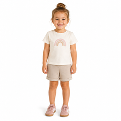 A young girl with her hair in a bun stands against a white background, wearing a cream-colored t-shirt with a pastel pink rainbow graphic, beige ribbed shorts, and pink trainers..