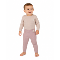 A smiling toddler standing barefoot against a plain white background, wearing a light beige long-sleeve top with a chest pocket, and dusty pink leggings with matching hem tag. 
