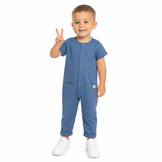 A toddler boy wearing a blue short-sleeve button-front jumpsuit with large front pockets, standing against a plain white background and making a peace sign with his hand.