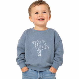 A young child wearing a blue/grey sweatshirt with a white graphic of a planet and astronaut, paired with blue jeans, photographed against a solid white background.