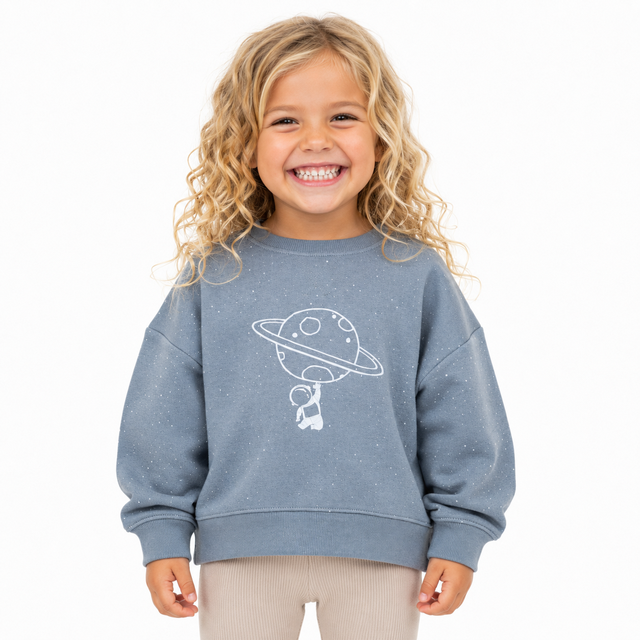 A young girl with curly blonde hair smiling, wearing a dusty grey/blue sweatshirt with a white line art graphic of a planet and astronaut, and light beige pants, against a solid white background.