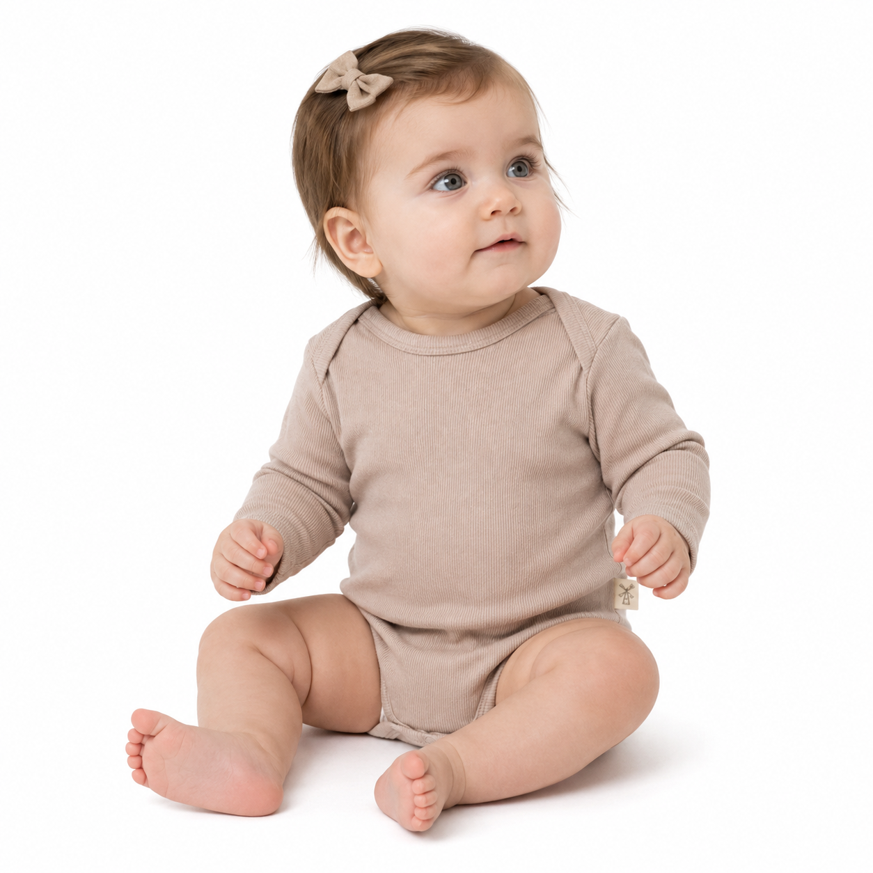 A baby girl sitting on a white background wearing a light beige long-sleeve ribbed bodysuit and a matching bow headband.