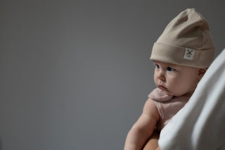 Baby being held and looking off to the side, wearing beige beanie hat and pink sleeveless romper