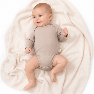 A baby lying on a cream-colored textured blanket, wearing a beige long-sleeve bodysuit. The baby is photographed from a top-down angle against a clean white background.