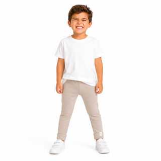 A young boy smiling and posing in a studio setting, wearing a plain white short-sleeve t-shirt, light beige slim-fit pants, and white trainers against a solid white background.