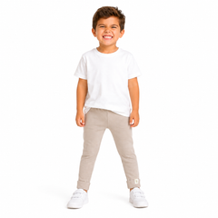 A young boy smiling and posing in a studio setting, wearing a plain white short-sleeve t-shirt, light beige slim-fit pants, and white trainers against a solid white background.