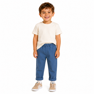 A young boy smiling, wearing a cream t-shirt, blue chinos, and beige sneakers, standing against a solid white background.