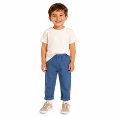 A young boy smiling, wearing a cream t-shirt, blue chinos, and beige sneakers, standing against a solid white background.