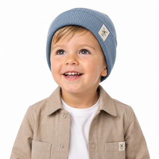 A smiling young child wearing a light blue ribbed knit beanie with a small white logo patch, paired with a beige shacket, over a white t-shirt, photographed against a solid white background.