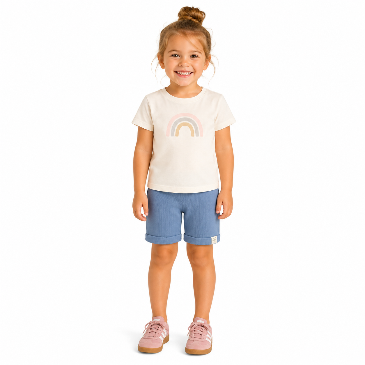 A young girl with her hair in a bun stands against a white background, wearing a cream-colored t-shirt with a pastel rainbow graphic, blue shorts, and pink trainers.