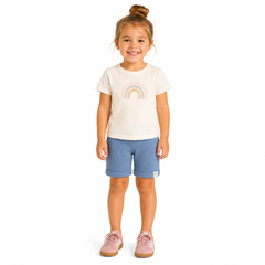 A young girl with her hair in a bun stands against a white background, wearing a cream-colored t-shirt with a pastel rainbow graphic, blue shorts, and pink trainers.