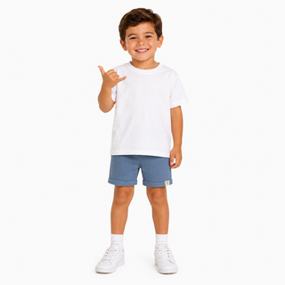 A young boy smiling and posing in a white short-sleeve t-shirt, blue shorts, white socks, and white trainers against a solid white background.