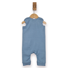 A baby sleeveless romper in dusty blue, shown from the back hanging on a wooden hanger from a wall hook against a white background.